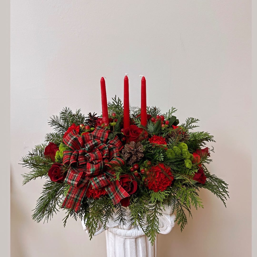 Red floral centerpiece with candles and a plaid bow in a white pedestal vase