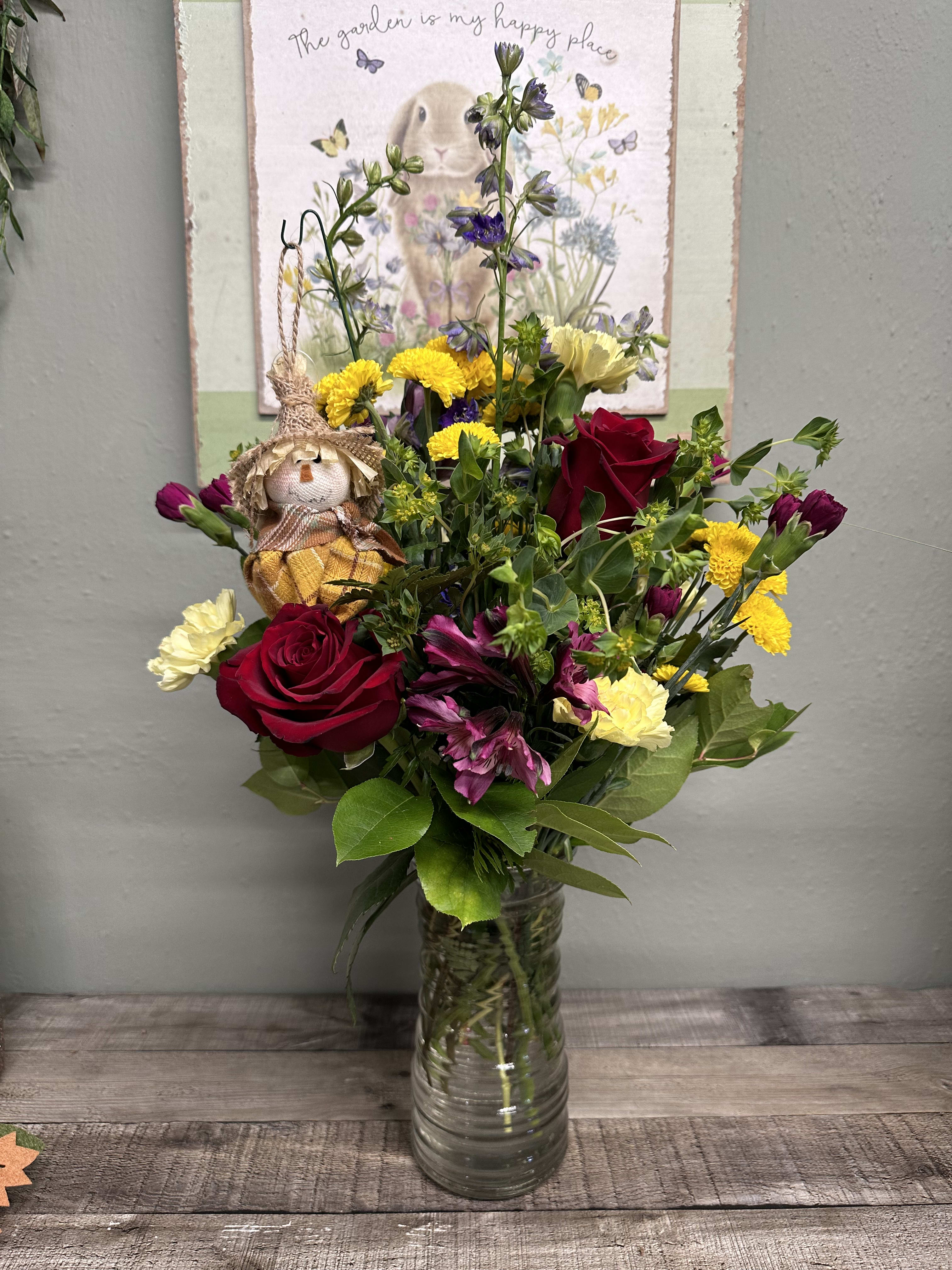 Mixed bouquet of red roses, yellow blooms, and purple flowers in a glass vase with a small scarecrow pick