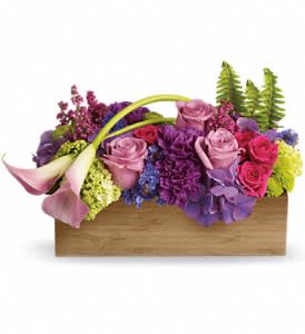 Low rectangular arrangement of pink and purple flowers with calla lilies and ferns in a wooden box.
