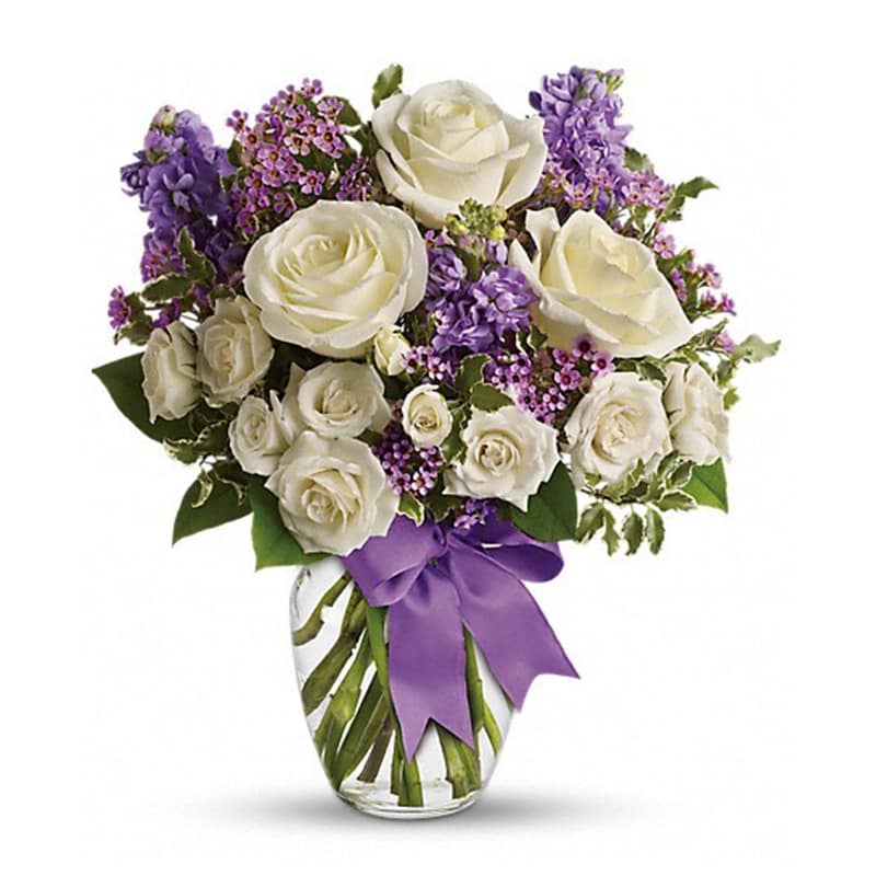 White roses and purple flowers arranged in a glass vase with a purple ribbon.