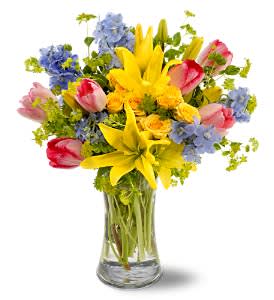 Mixed bouquet of yellow lilies, pink tulips, and blue flowers in a glass vase