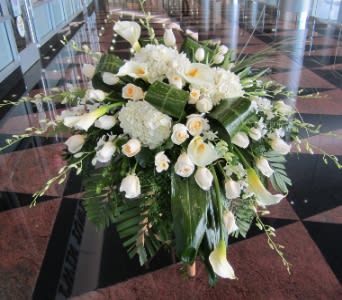 Large white arrangement of roses, calla lilies, and hydrangeas with bold green leaves on a stand.