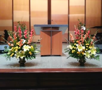 Pair of tall yellow and white lily arrangements with pink blooms on urns flanking a wooden podium