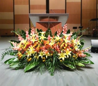 Low, wide arrangement of yellow lilies and peach flowers with large green foliage in front of a stage podium