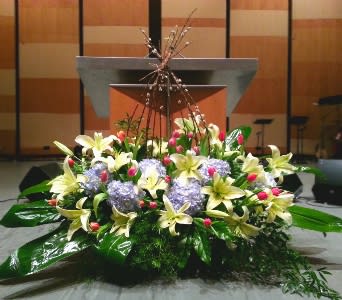 Low altar arrangement of yellow lilies, lavender hydrangeas, and pink buds with large glossy foliage.