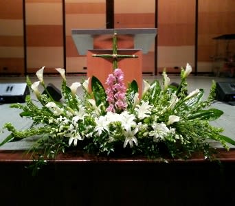 Large white and green altar arrangement with lilies, calla lilies, and a pink flower cluster beneath a green cross.