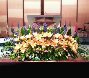 Low altar spray of peach lilies, blue flowers, and pink buds in a lush horizontal display.