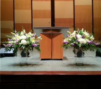 Two large white and purple floral arrangements in tall clear vases flanking a wooden podium