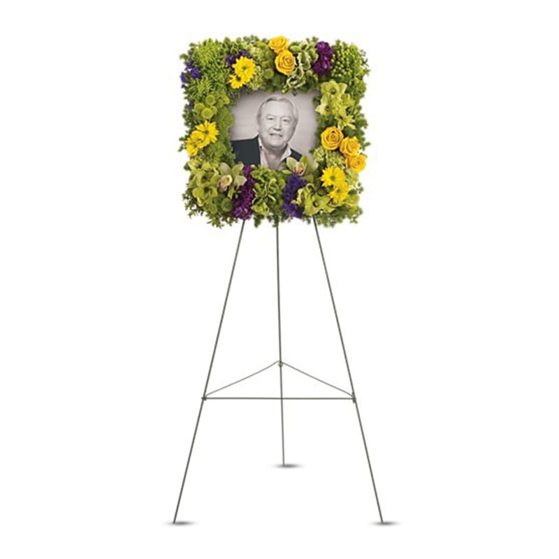 Tall easel-mounted square floral frame of green, yellow, and purple flowers surrounding a black-and-white photo.