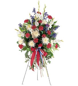 Standing spray of red carnations, white mums, and blue flowers on an easel with a patriotic ribbon.