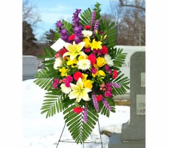 Standing spray of yellow lilies, bright pink flowers, and white mums on an easel in a cemetery