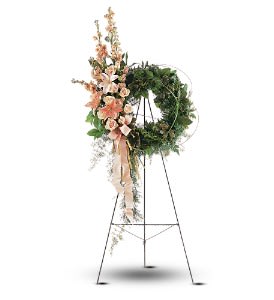 Standing green wreath with peach lilies and carnations on a tall metal easel with cascading ribbon.