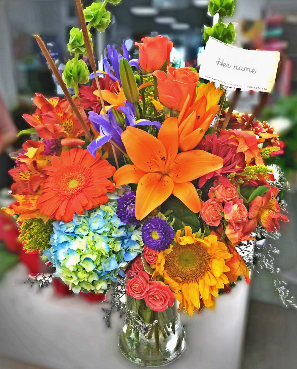 Bright mixed bouquet with orange lilies, roses, sunflowers, and blue hydrangea in a glass vase