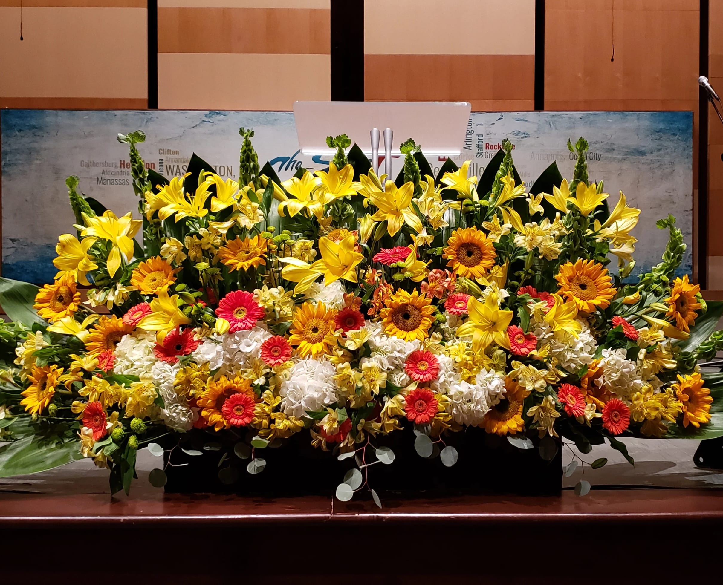Large stage arrangement of yellow lilies, orange gerbera daisies, and white hydrangeas in a low black container