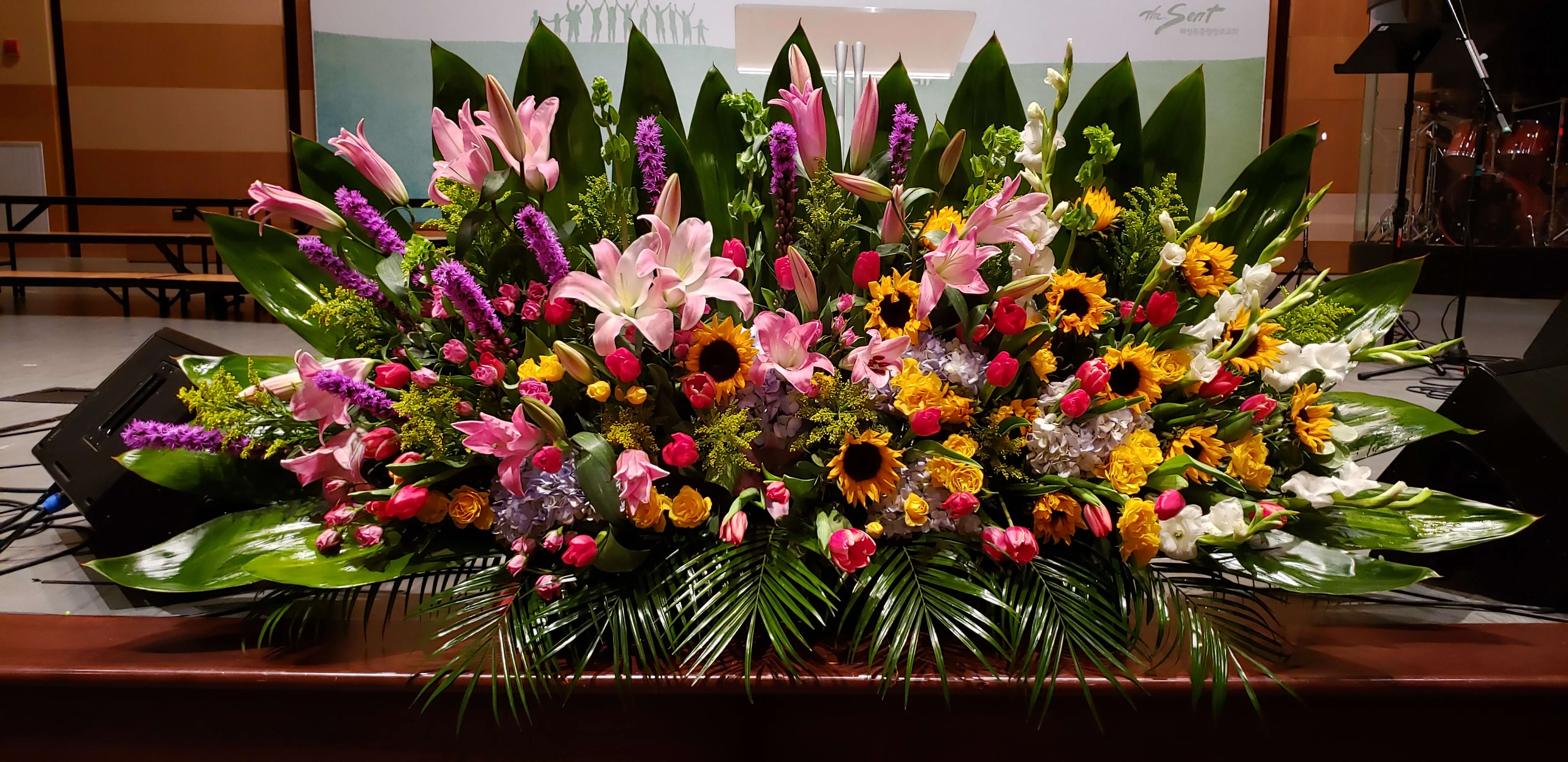 Large horizontal event arrangement of pink lilies, tulips, sunflowers, and mixed blooms with bold tropical foliage