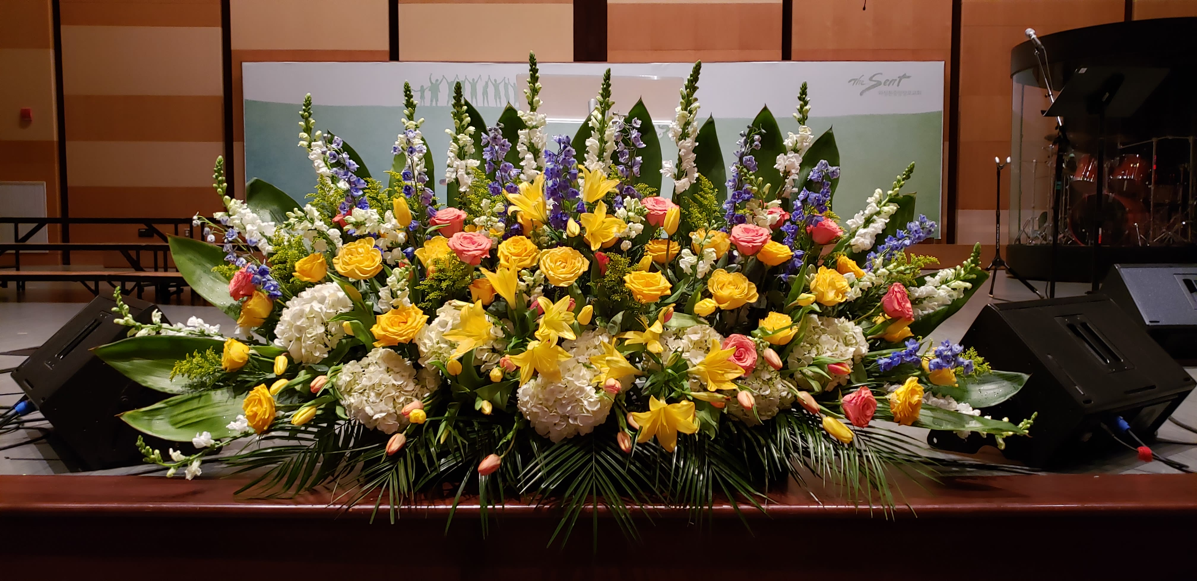 Large stage arrangement of yellow lilies, roses, white hydrangea, and blue delphinium.