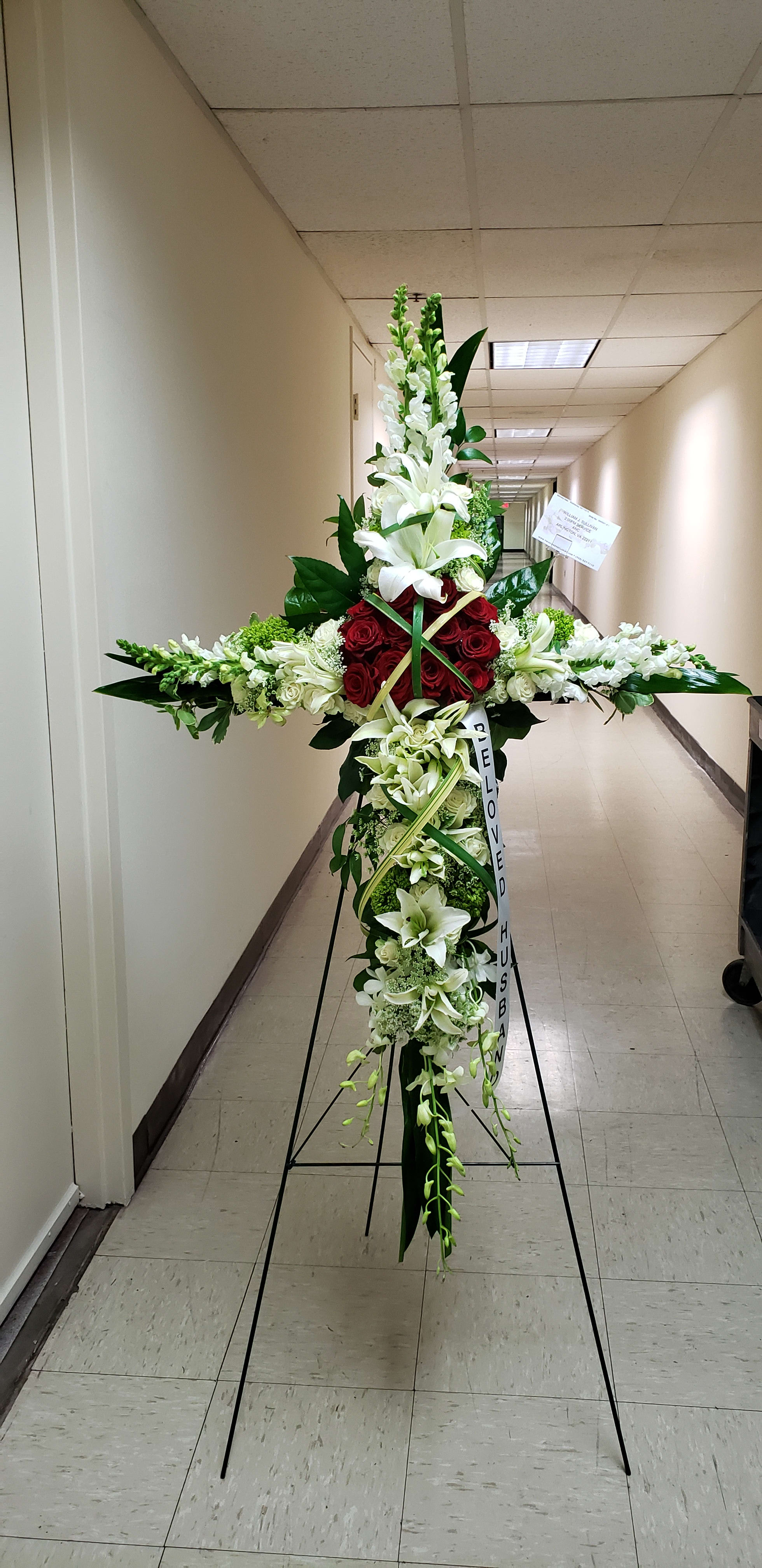 Tall cross-shaped funeral spray with white lilies, snapdragons, and red roses on a metal easel