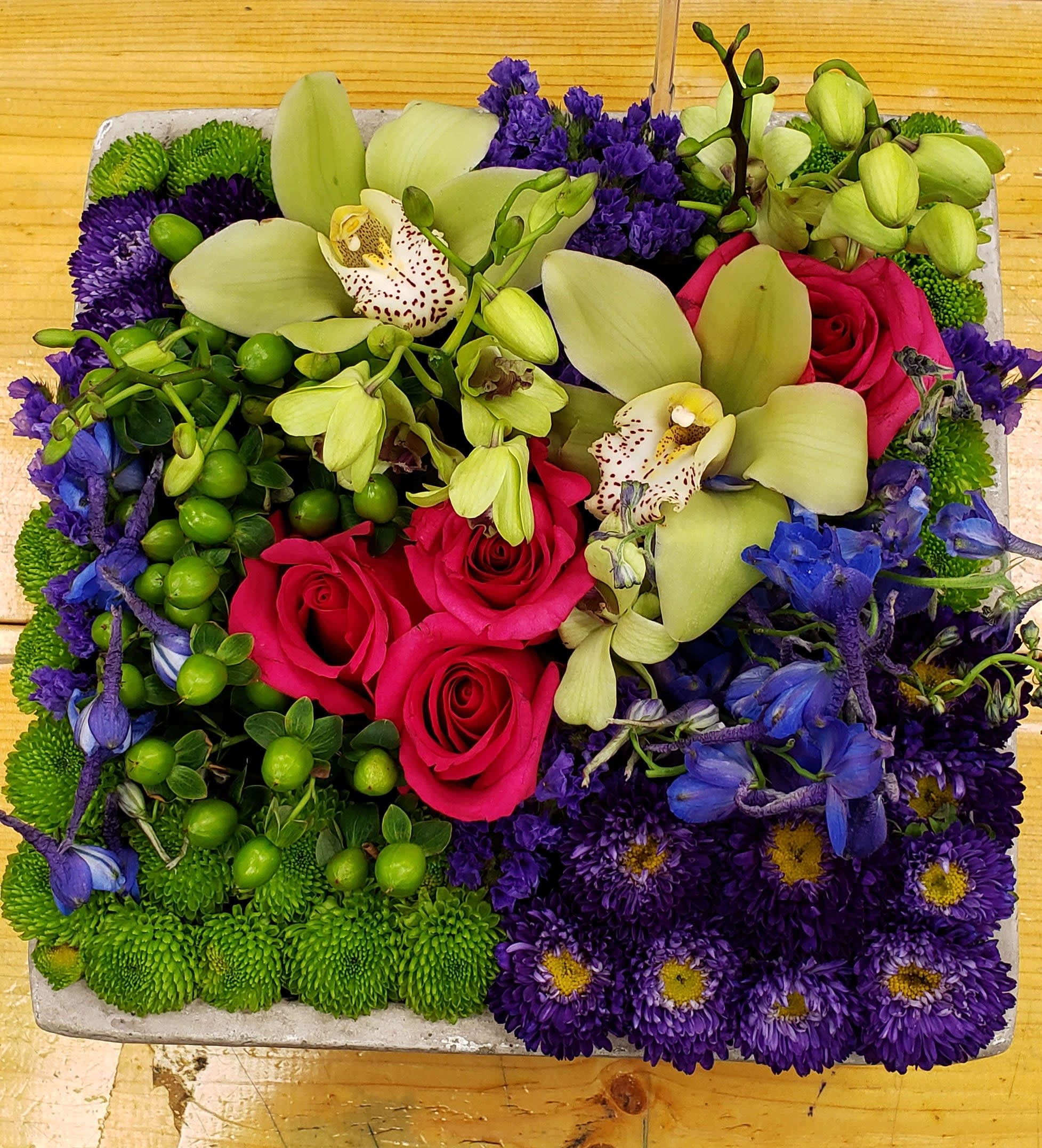 Square arrangement with pink roses, green orchids, purple mums and blue delphinium in a low container