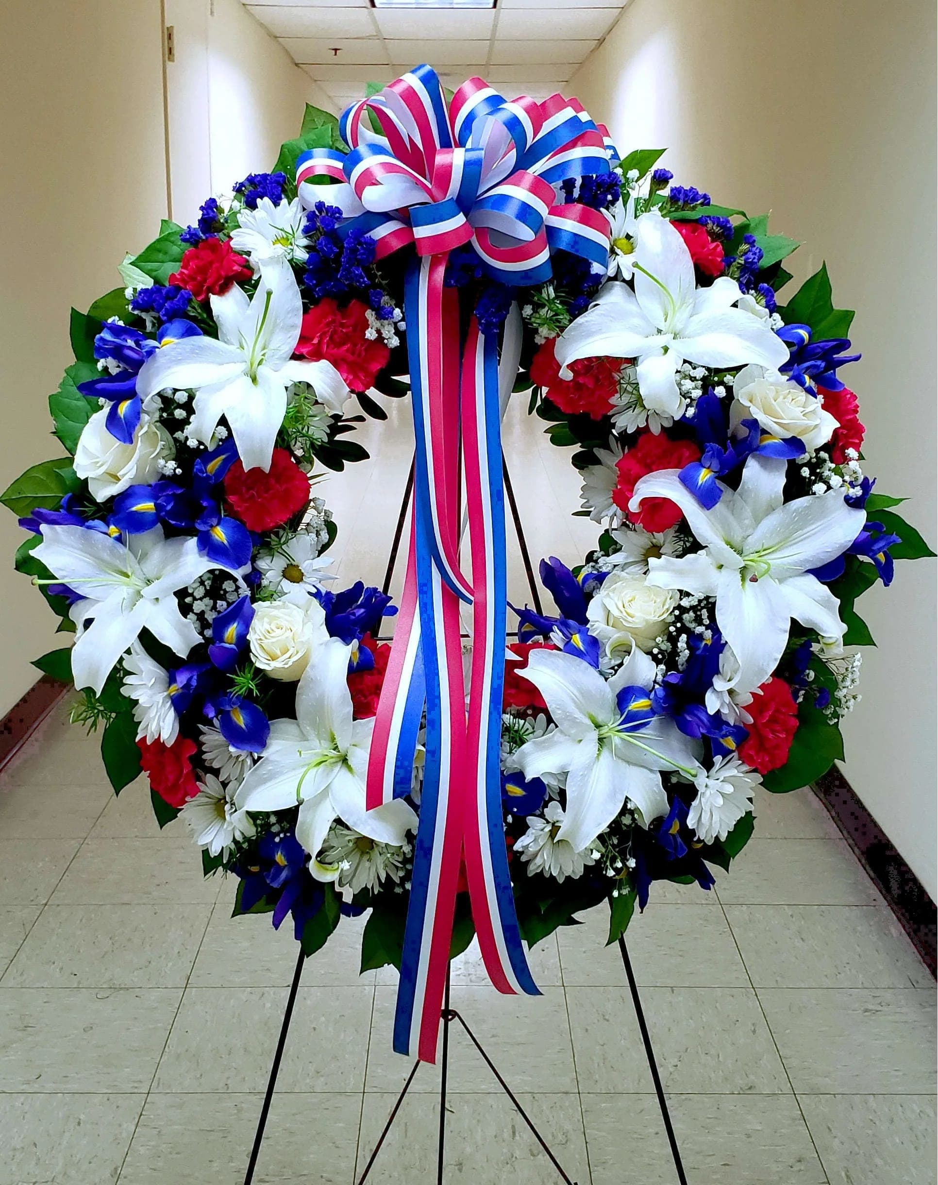 Large floral wreath with white lilies, red carnations, blue flowers, and a tricolor ribbon bow