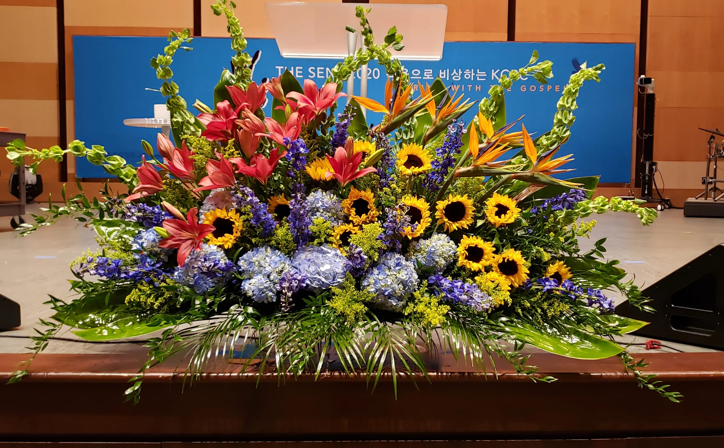 Large stage arrangement with yellow sunflowers, pink lilies, blue hydrangeas and orange tropical flowers.