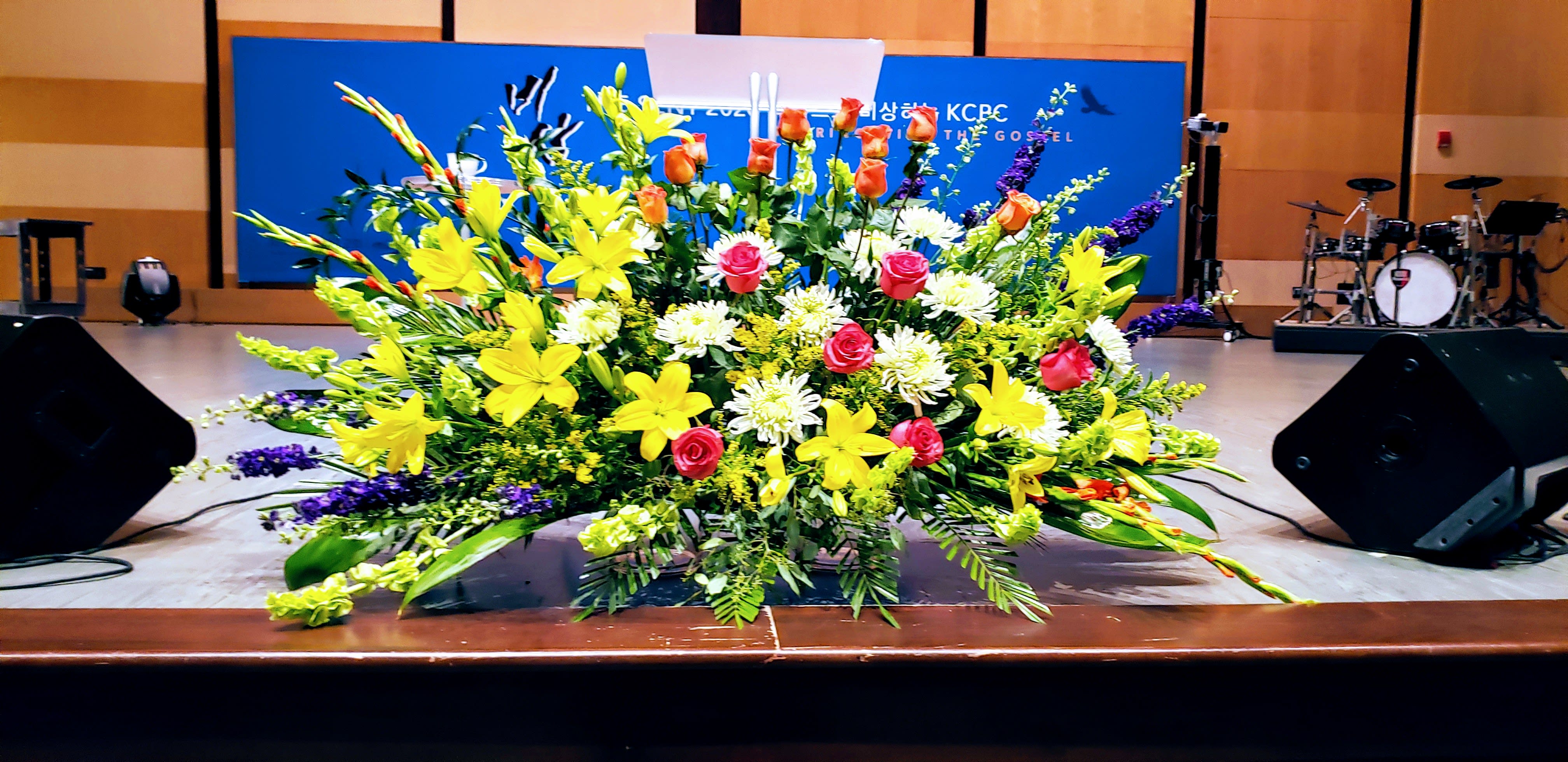 Large altar-style arrangement with yellow lilies, pink roses, and white mums on a stage