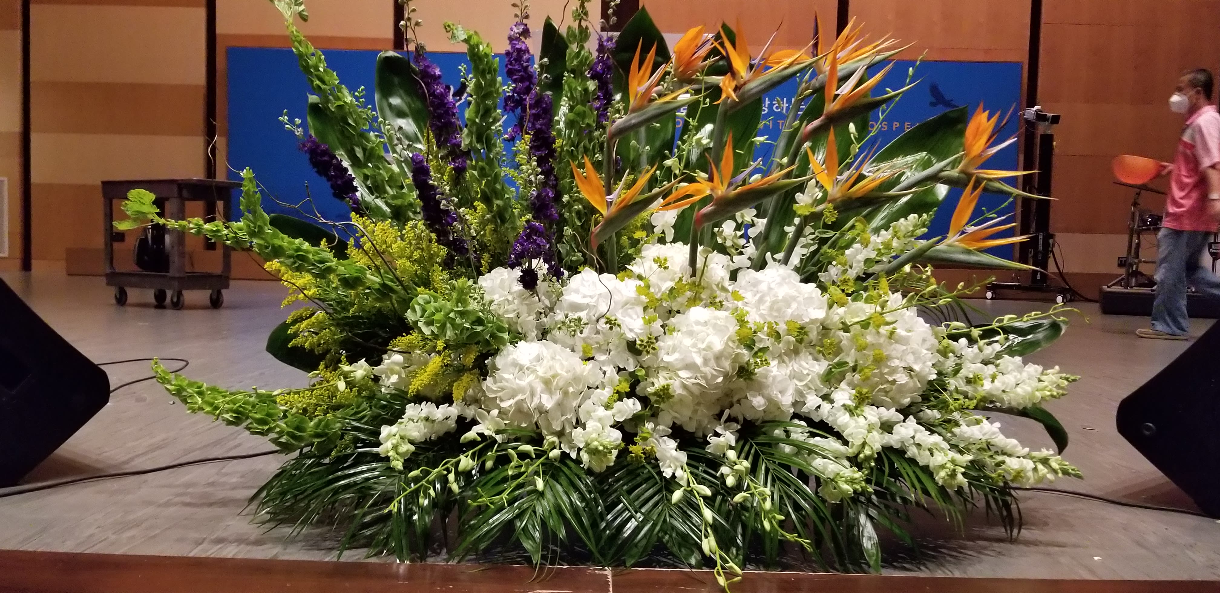 Large stage arrangement with white hydrangeas, orange tropical blooms, and tall green and purple accents