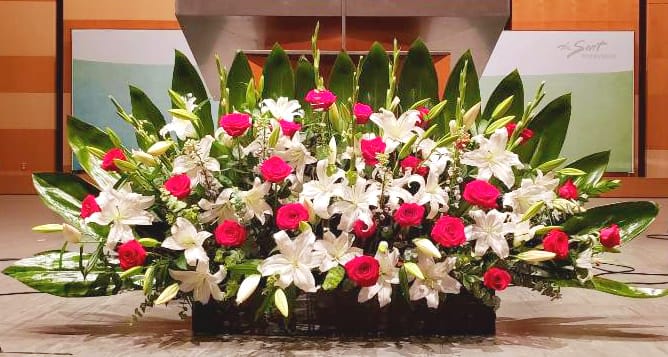 Large low arrangement of red roses and white lilies with bold green leaves on a stage.