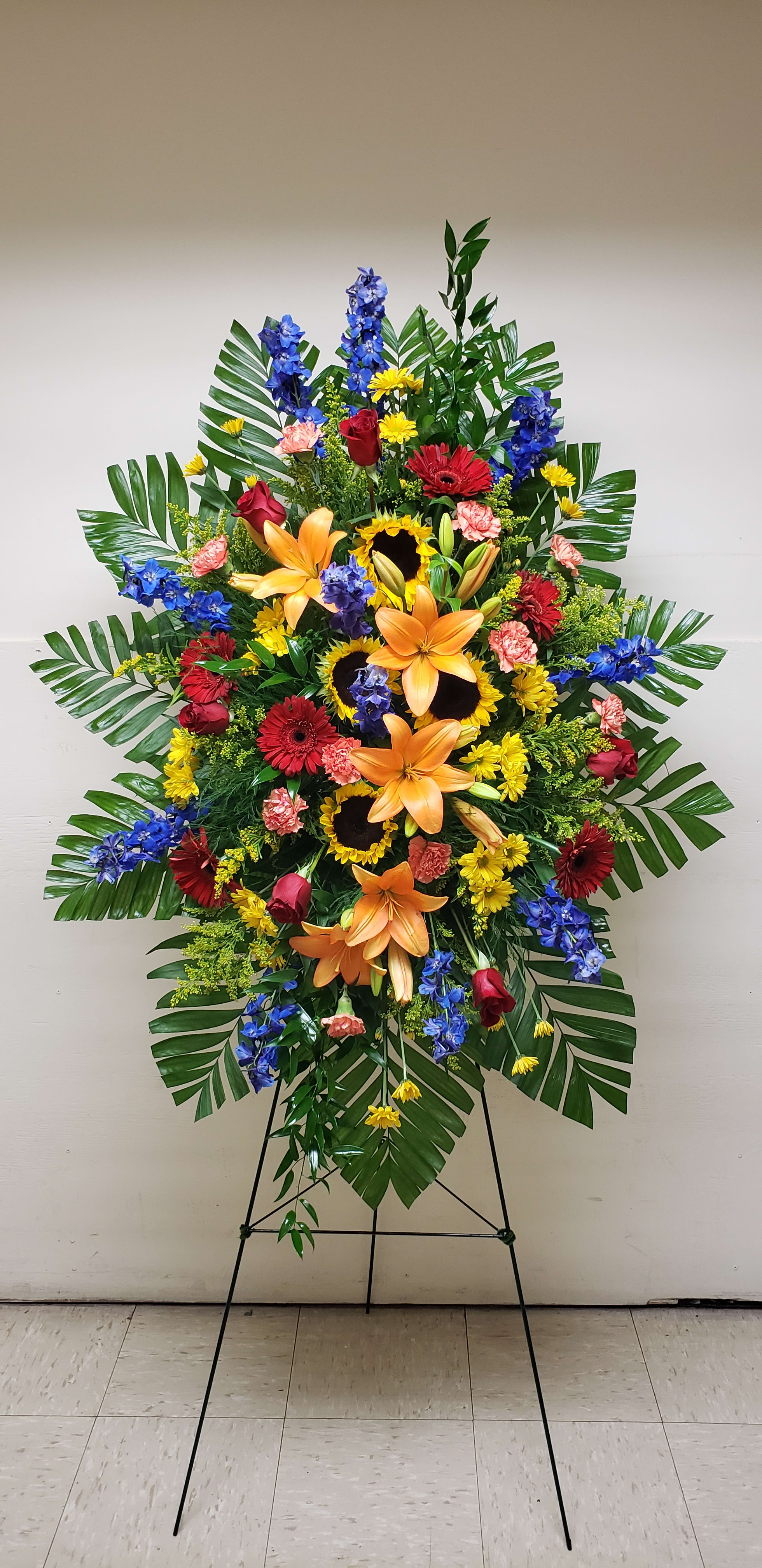 Tall standing spray of orange lilies, sunflowers, red roses, and mixed bright flowers on an easel.