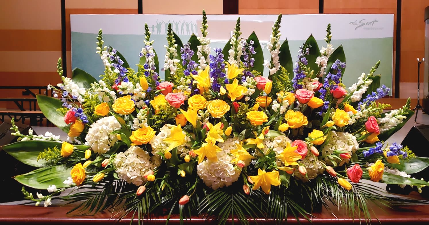 Large altar flower arrangement with yellow lilies and roses, white hydrangeas, pink roses, and blue delphinium.