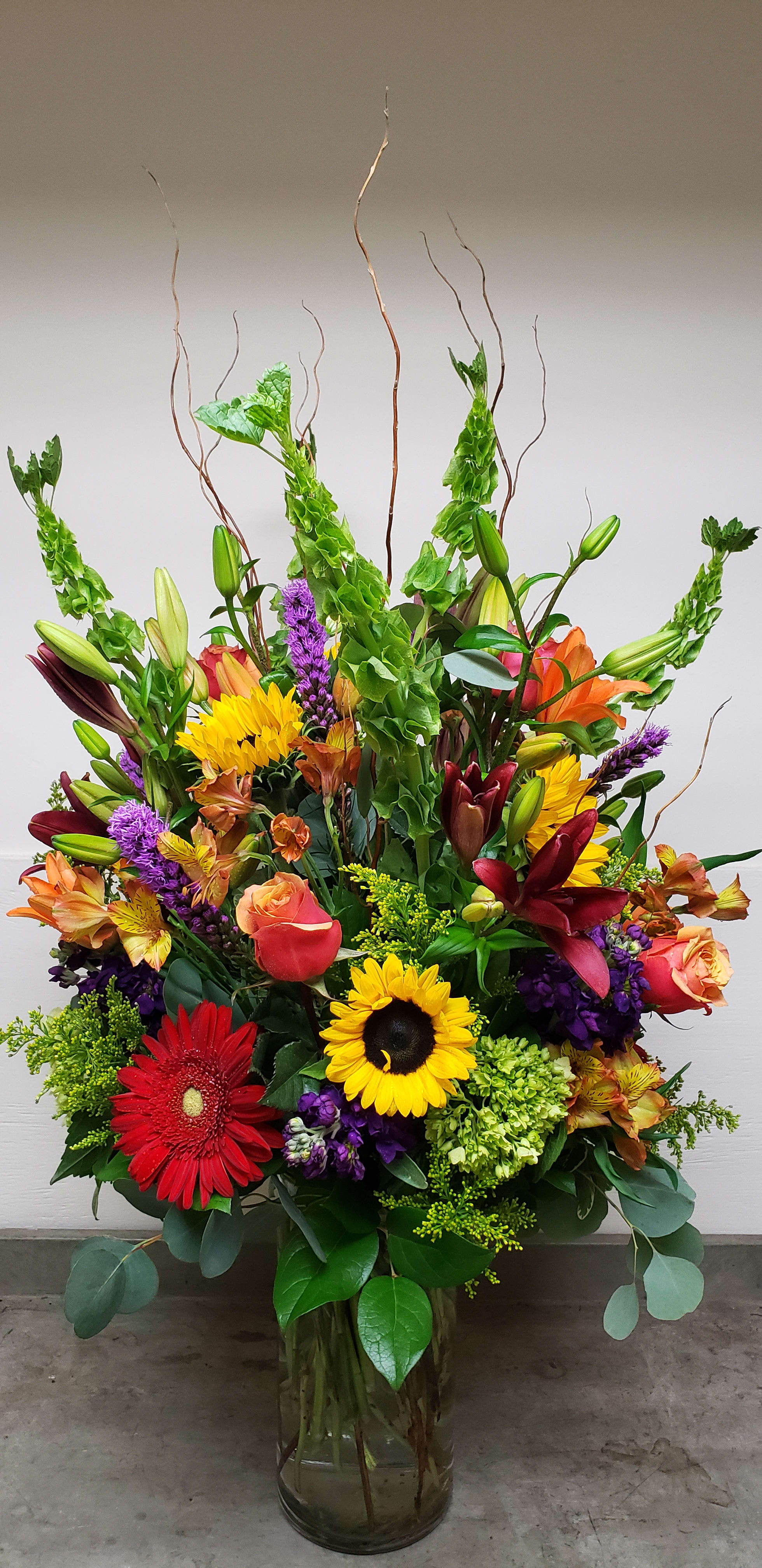 Tall mixed arrangement with sunflowers, lilies, roses, gerbera and bells of Ireland in a clear glass vase