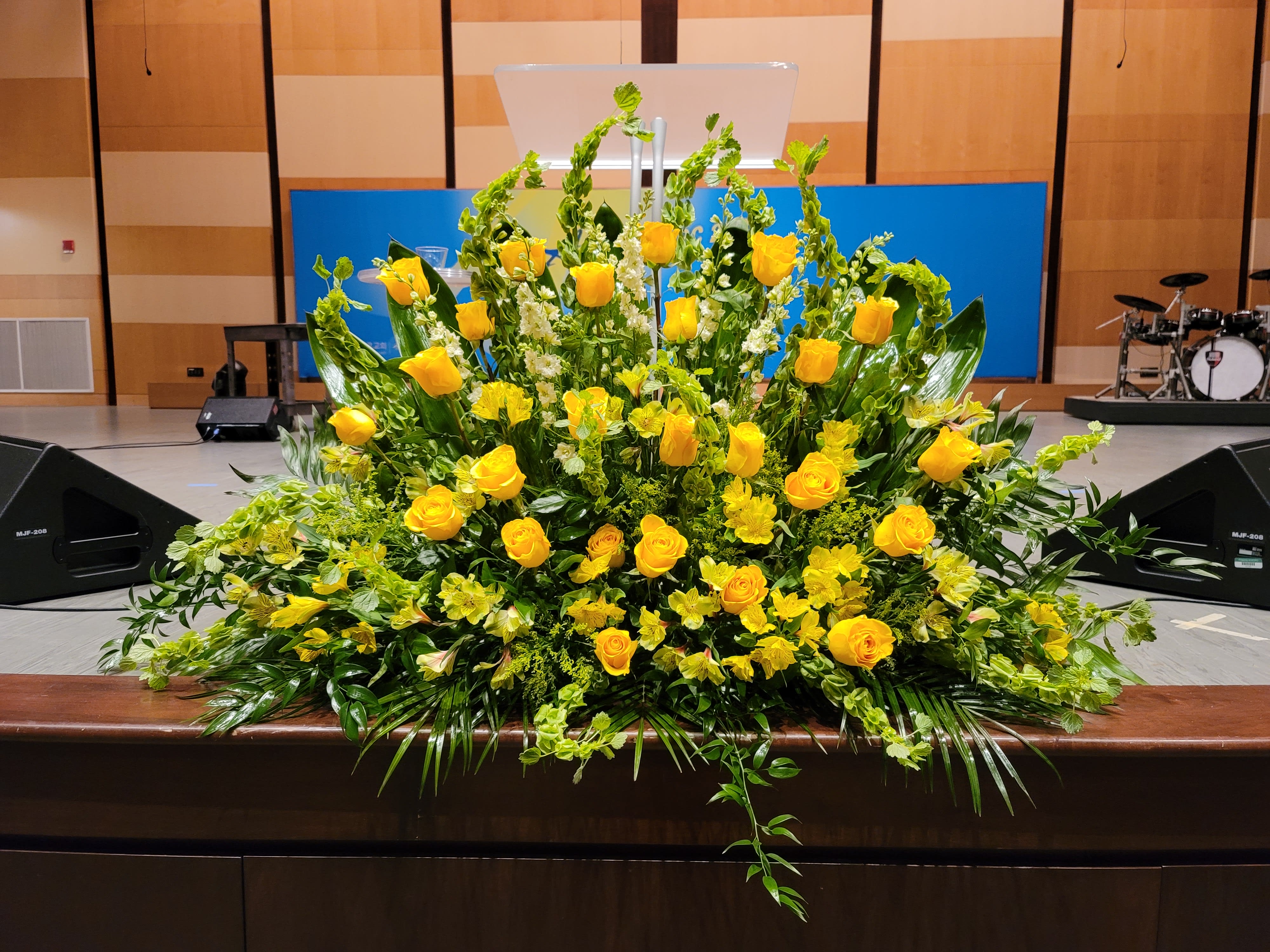 Large fan-shaped arrangement of yellow roses and flowers with lush greenery on a stage front.