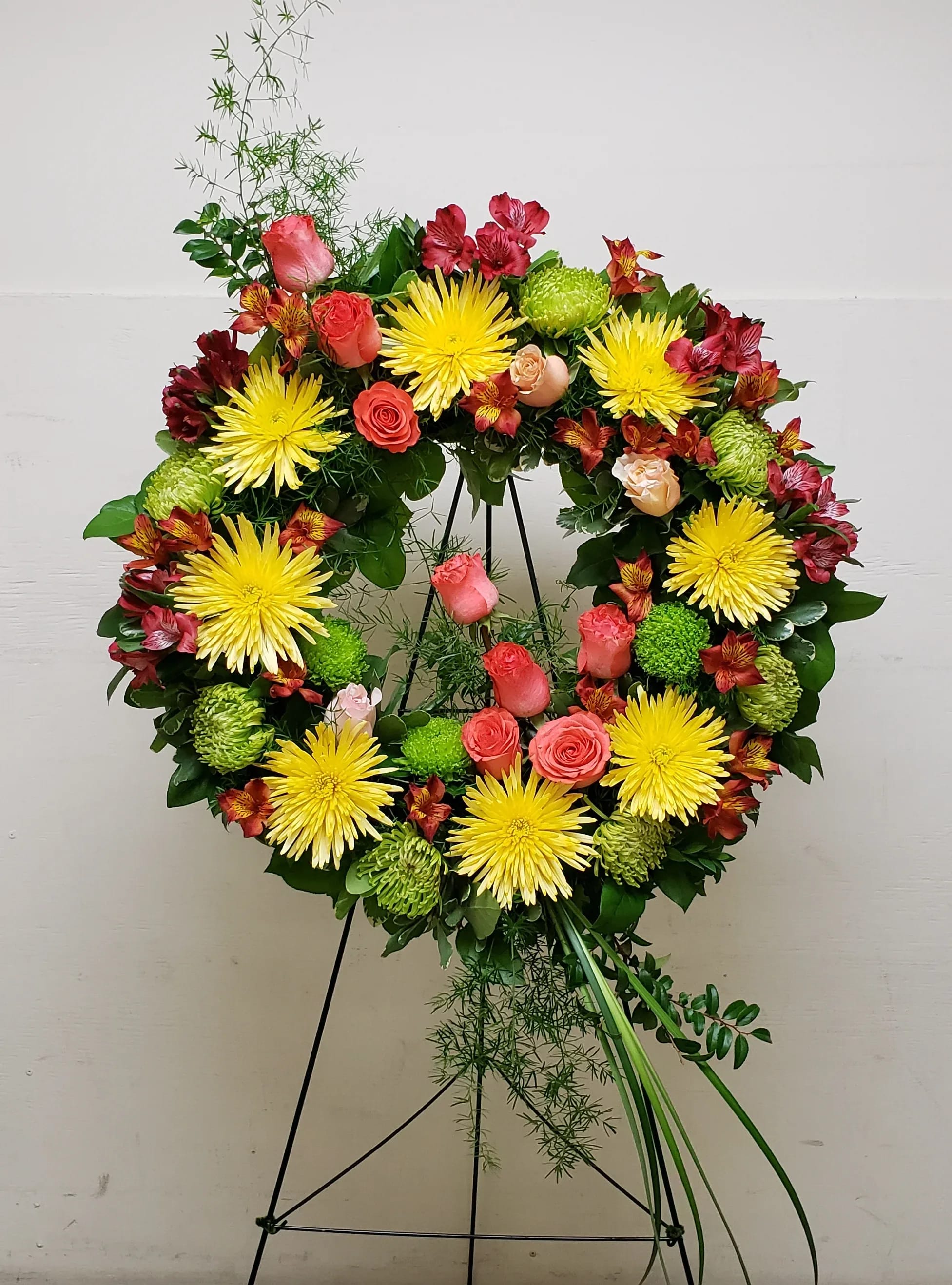 Standing wreath of yellow mums, pink roses, and red flowers on a green easel display