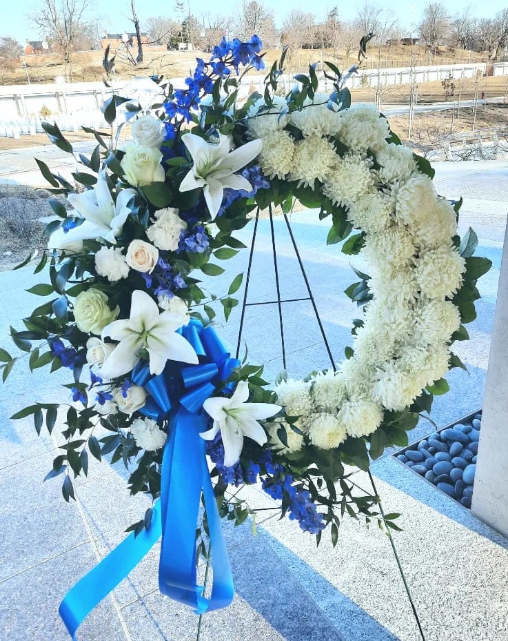 Standing white and blue funeral wreath with lilies and ribbon on a metal easel.