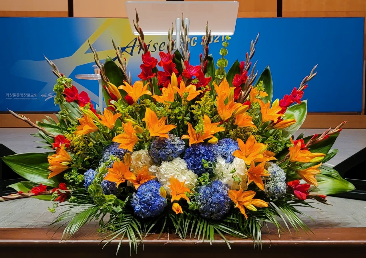 Large altar floral arrangement with orange lilies, blue and white hydrangeas, and red accent flowers on a stage