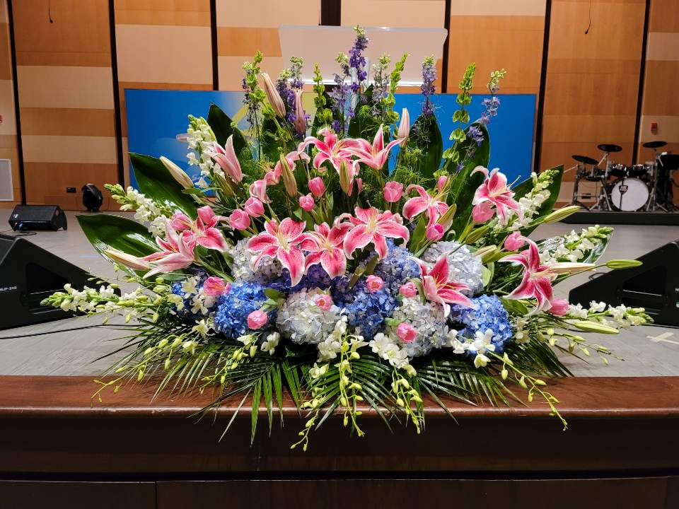 Large altar-style arrangement of pink lilies, blue hydrangeas, and white flowers in a fan shape