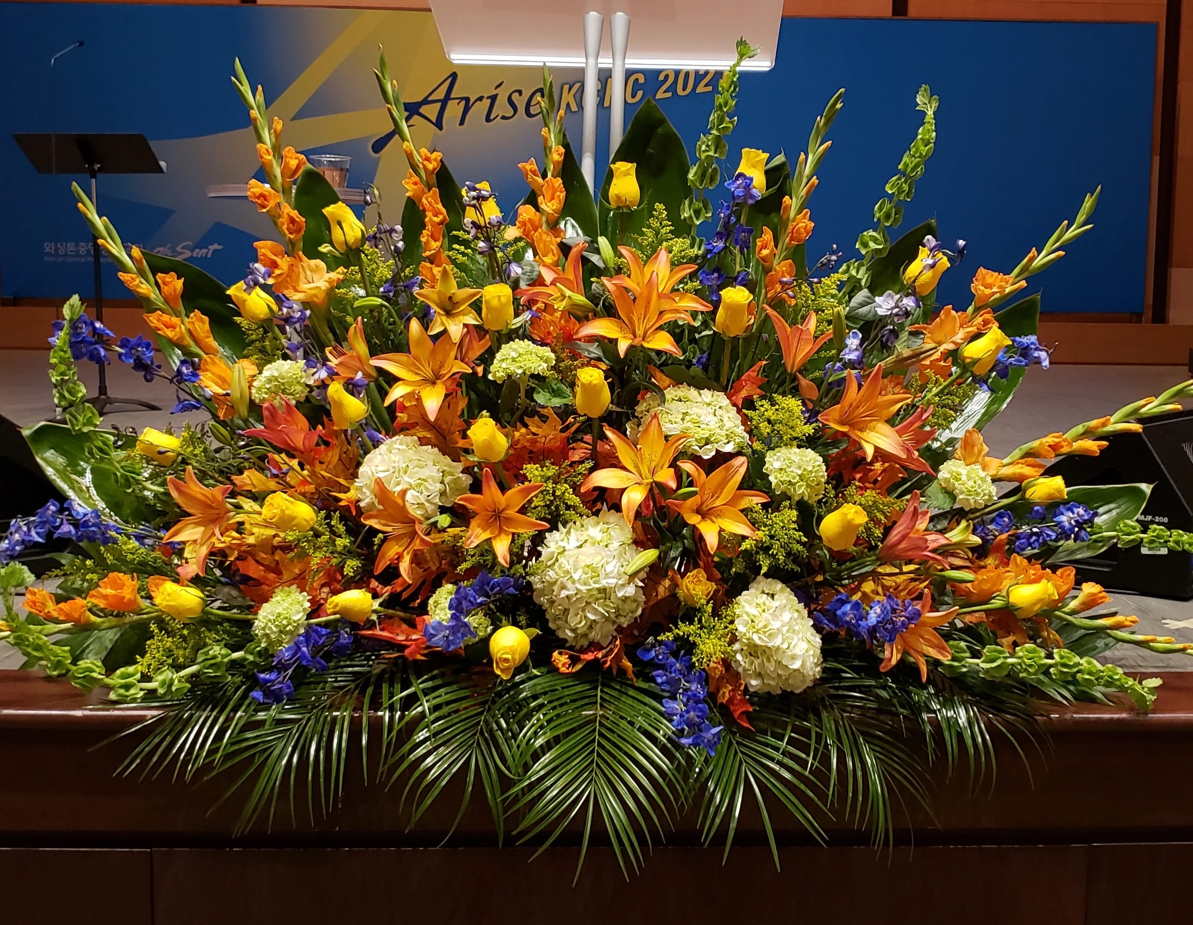 Large altar arrangement with orange lilies, yellow roses, blue delphinium, and white hydrangeas in a fan shape