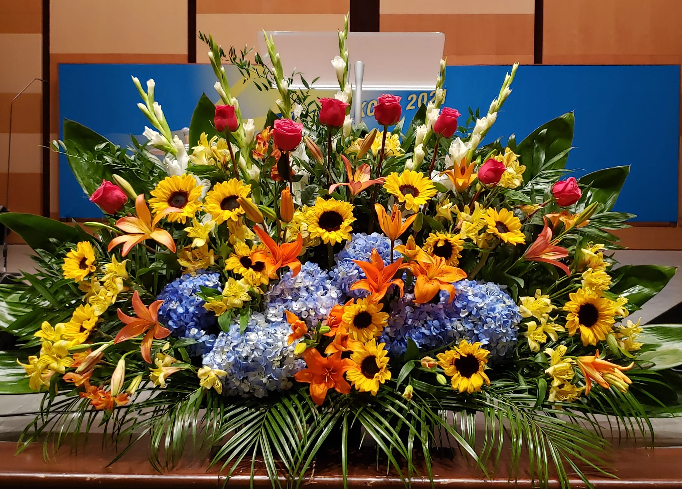 Large stage arrangement with yellow sunflowers, blue hydrangeas, orange lilies, and pink roses