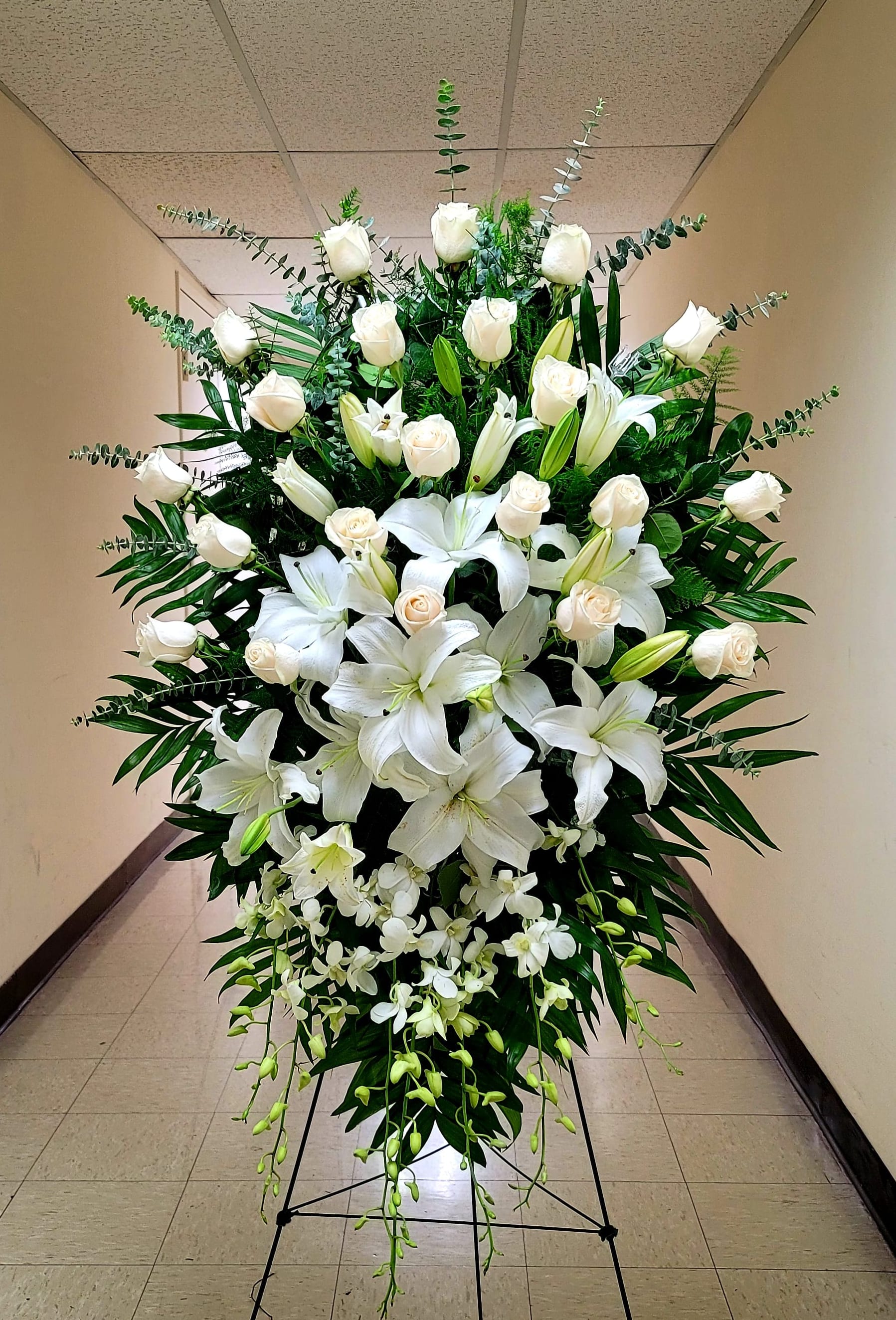Tall standing spray of white roses, lilies, and orchids on an easel.