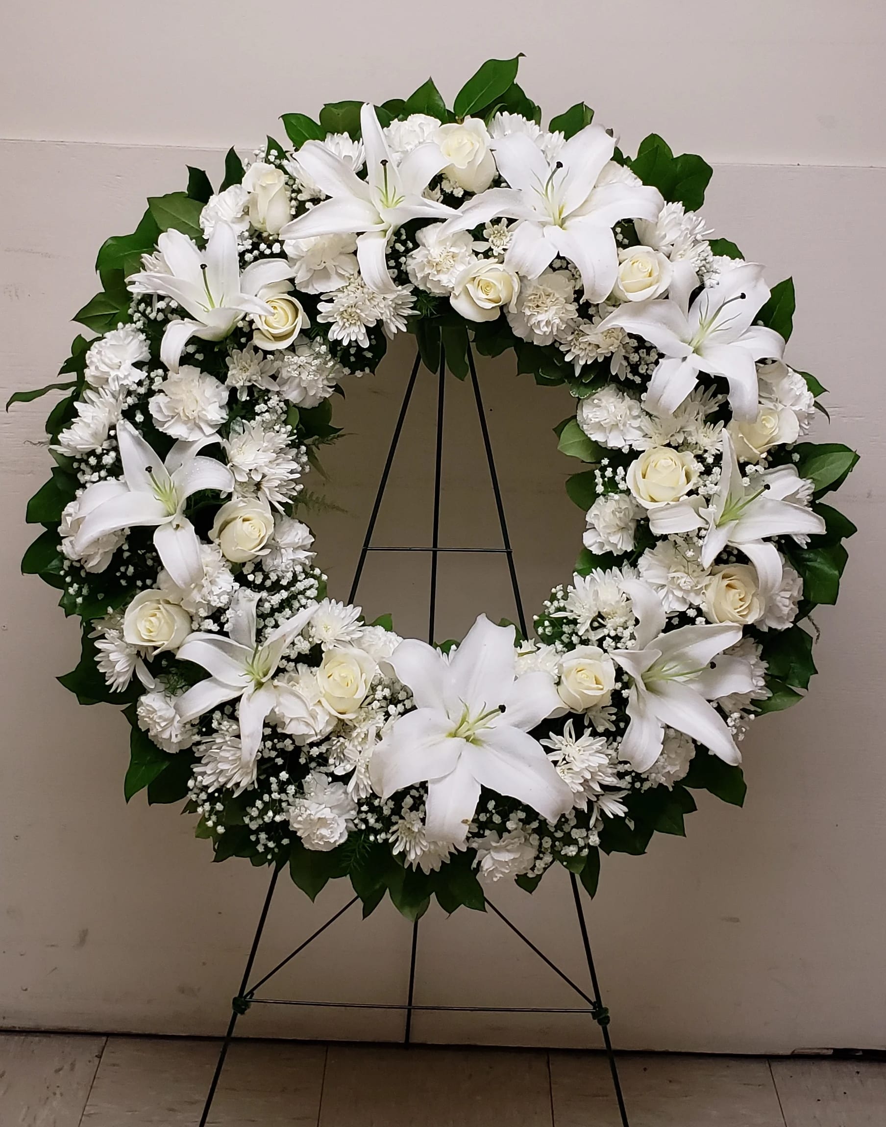 Large white floral wreath with lilies, roses, and mums displayed on a metal easel stand