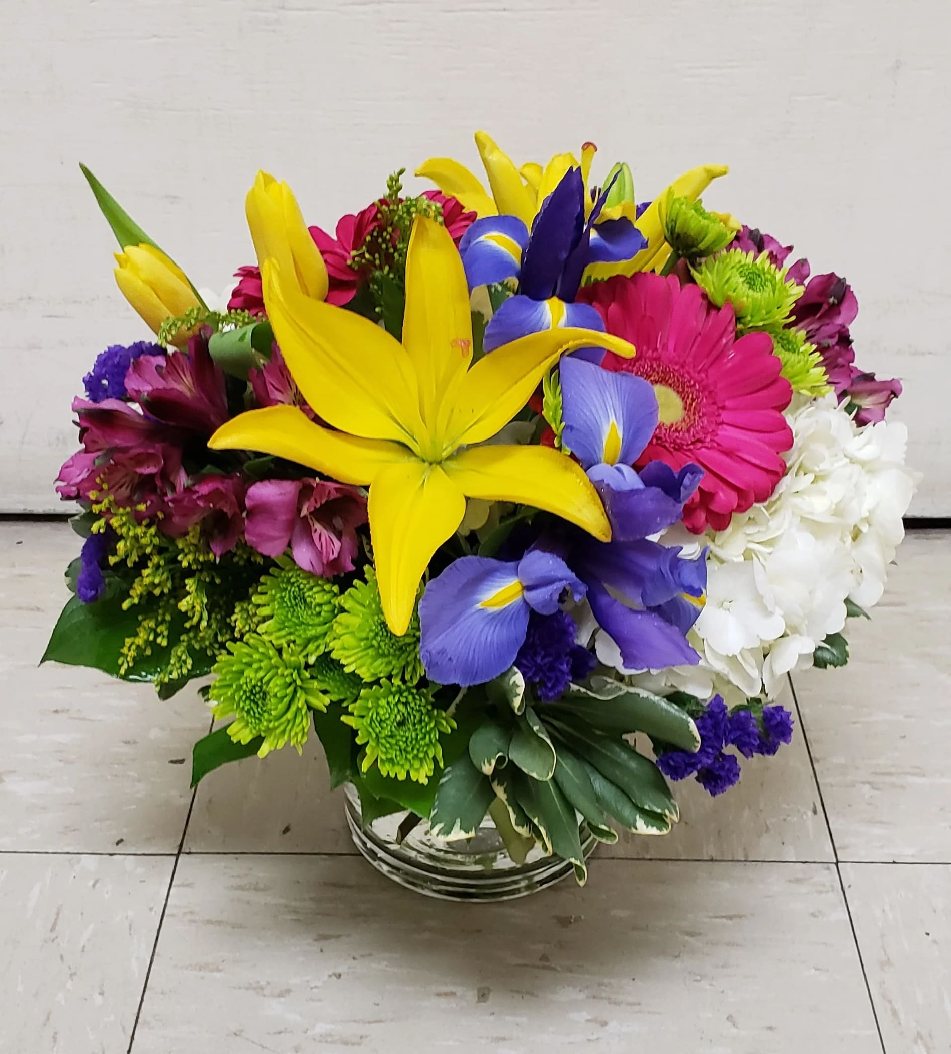 Bright mixed bouquet with yellow lilies, purple irises, pink gerberas, and white hydrangeas in a glass vase