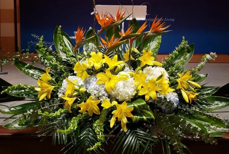Large altar-style arrangement with yellow lilies, white hydrangeas, and orange tropical flowers in lush foliage