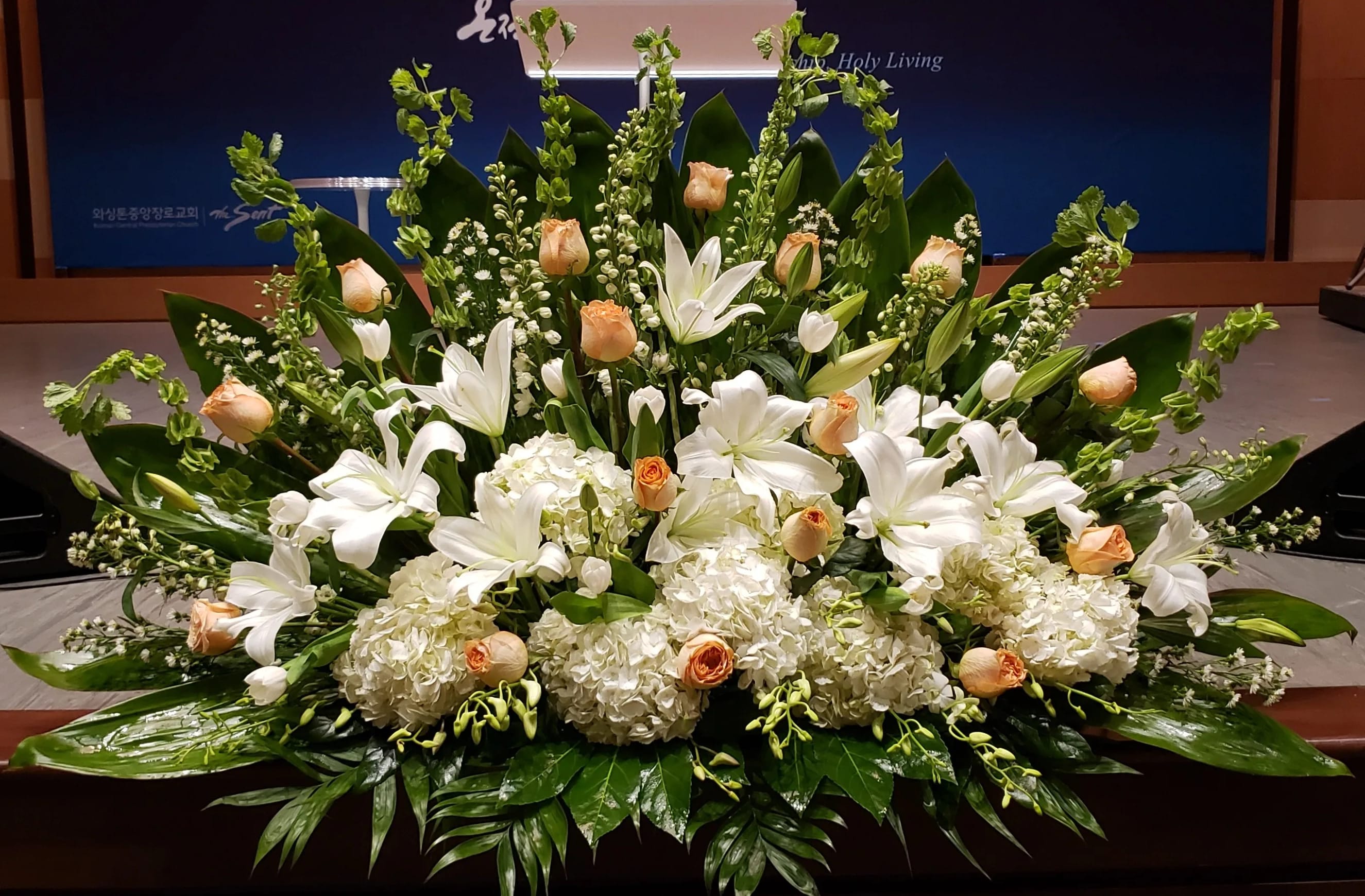 Low, wide arrangement of white lilies, hydrangeas, and peach roses on a stage