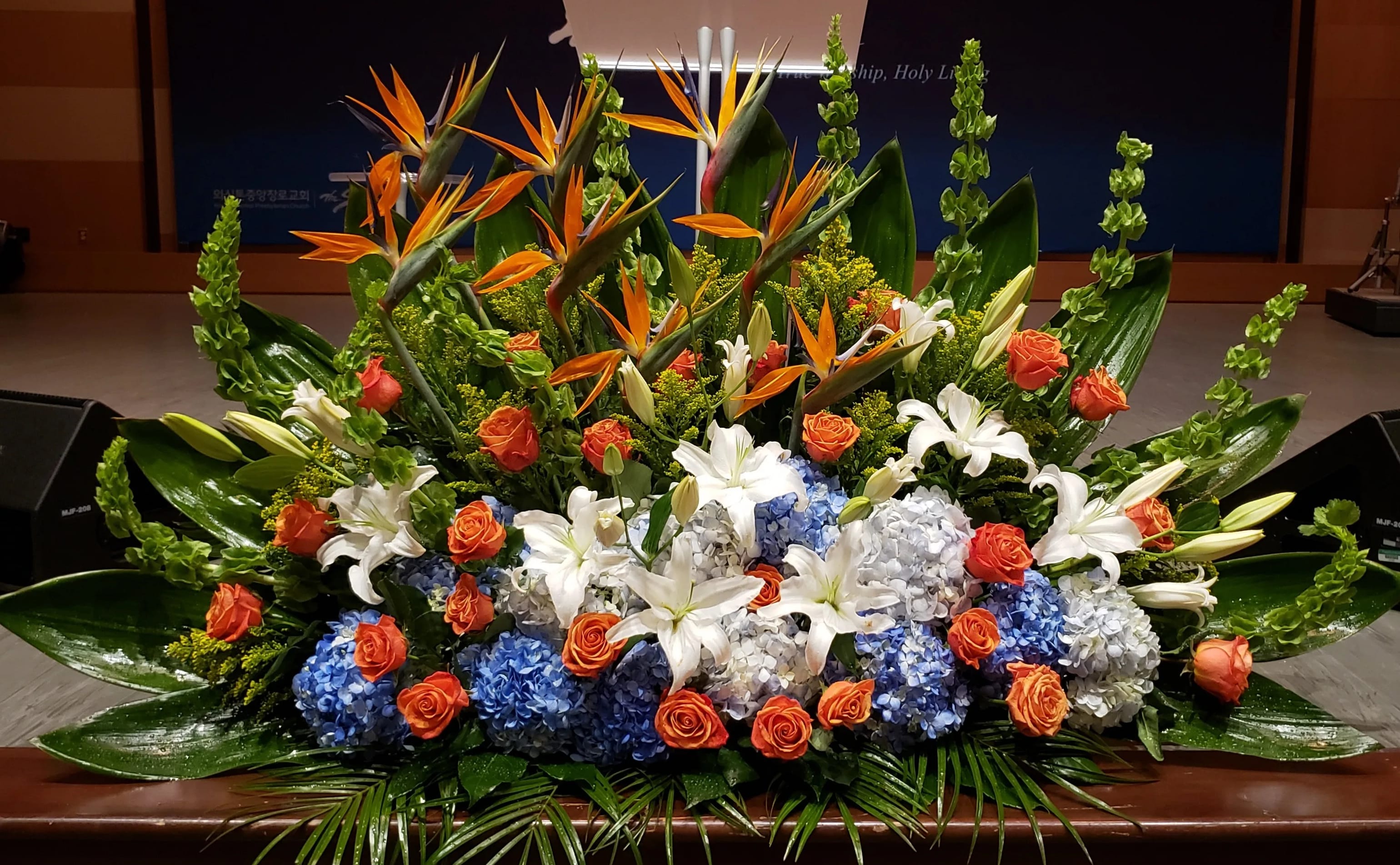 Large altar-style arrangement with orange, blue, and white flowers including lilies and hydrangeas