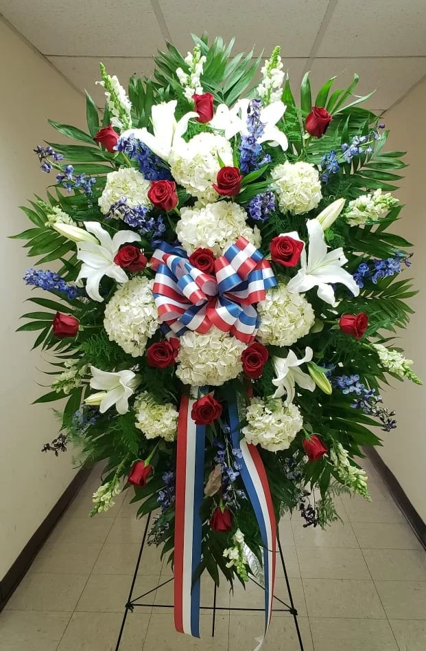 Tall standing spray of red roses, white lilies, white hydrangeas, and blue flowers with a red, white, and blue bow.
