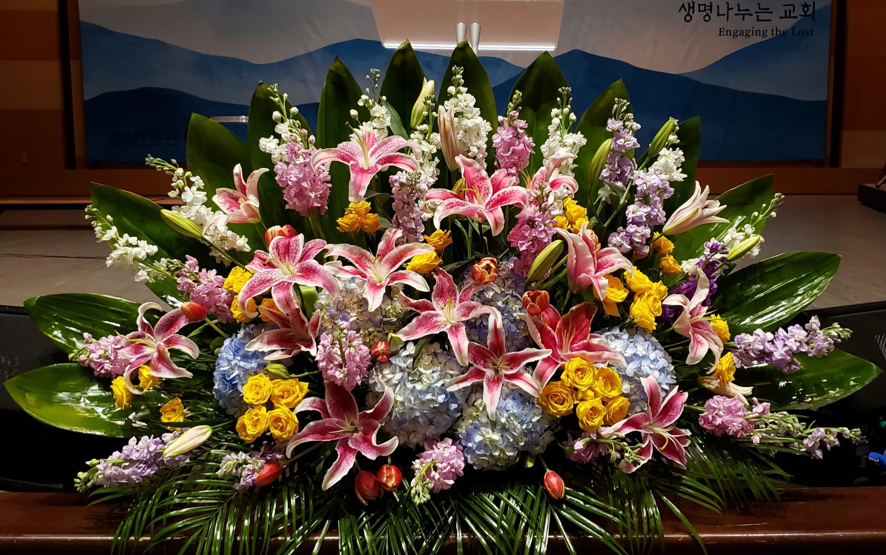 Large fan-shaped arrangement of pink lilies, yellow roses, blue hydrangeas and pastel blooms framed by glossy green leaves