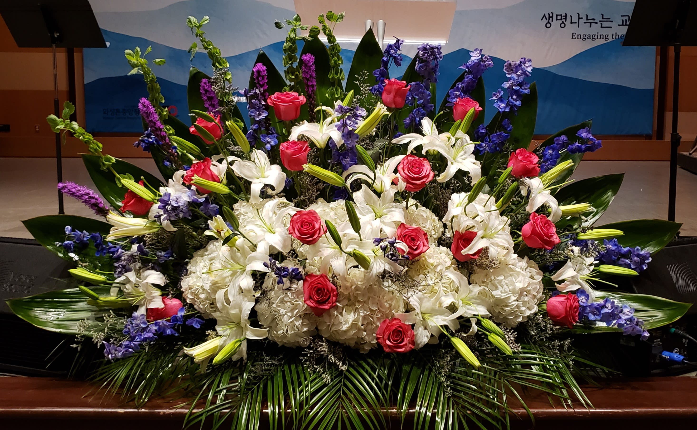 Large altar-style arrangement of white lilies and hydrangeas with red roses and blue delphinium in a fan shape
