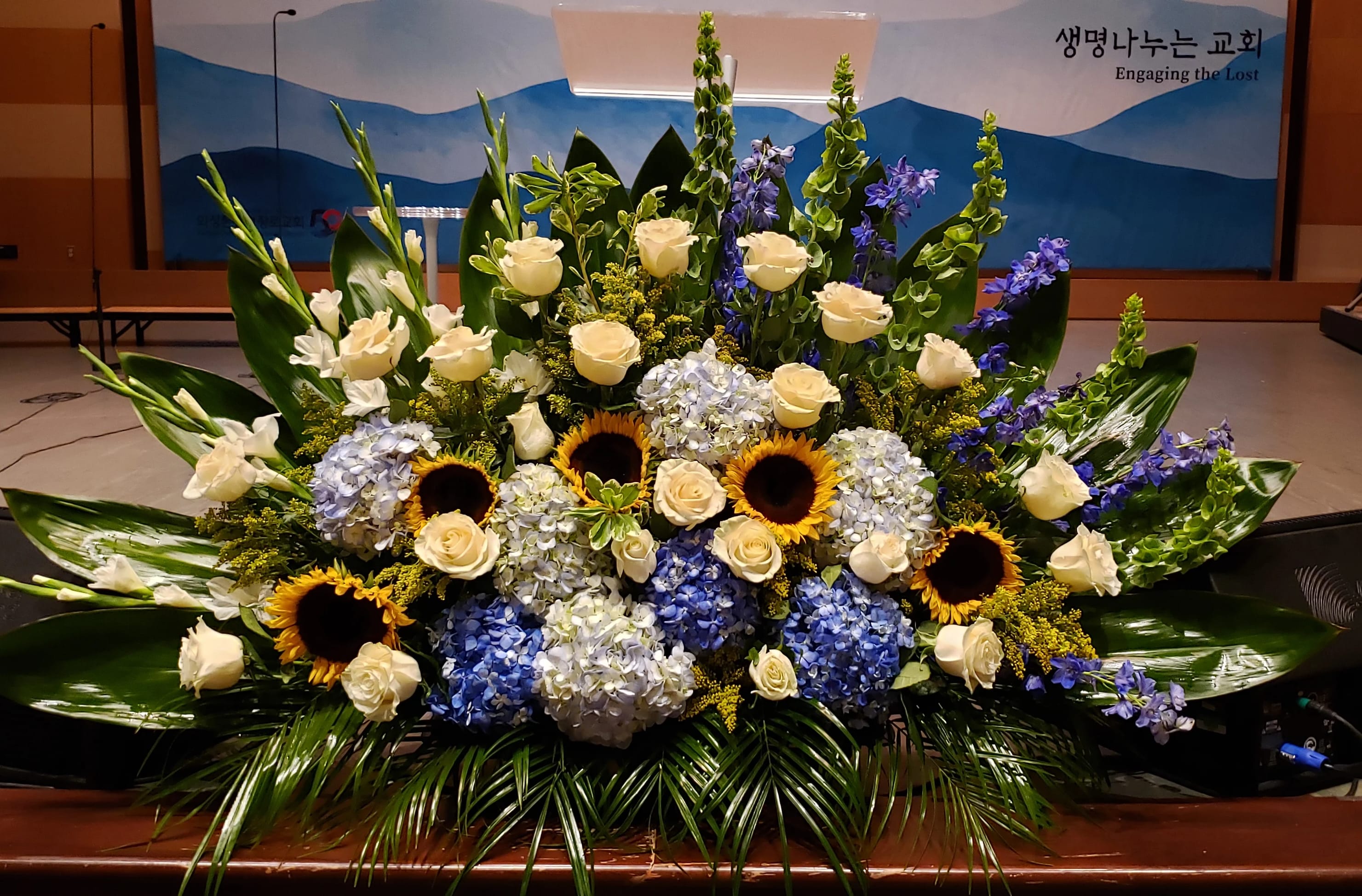 Large floral arrangement with white roses, blue hydrangeas, sunflowers, and purple spikes