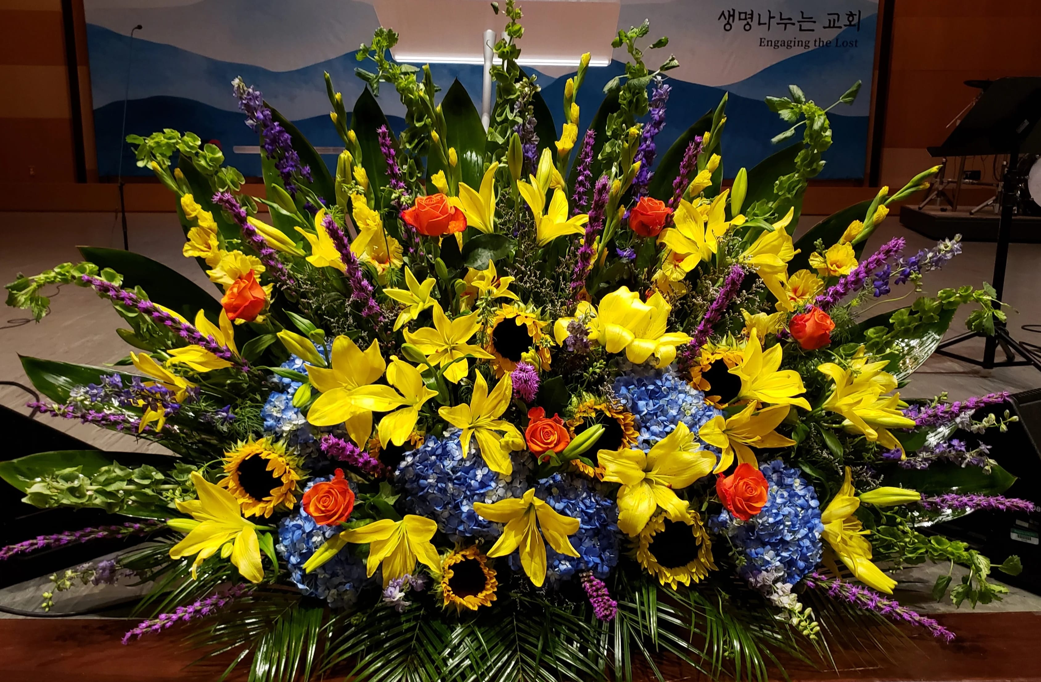 Large floral arrangement with yellow lilies, blue hydrangeas, and orange roses