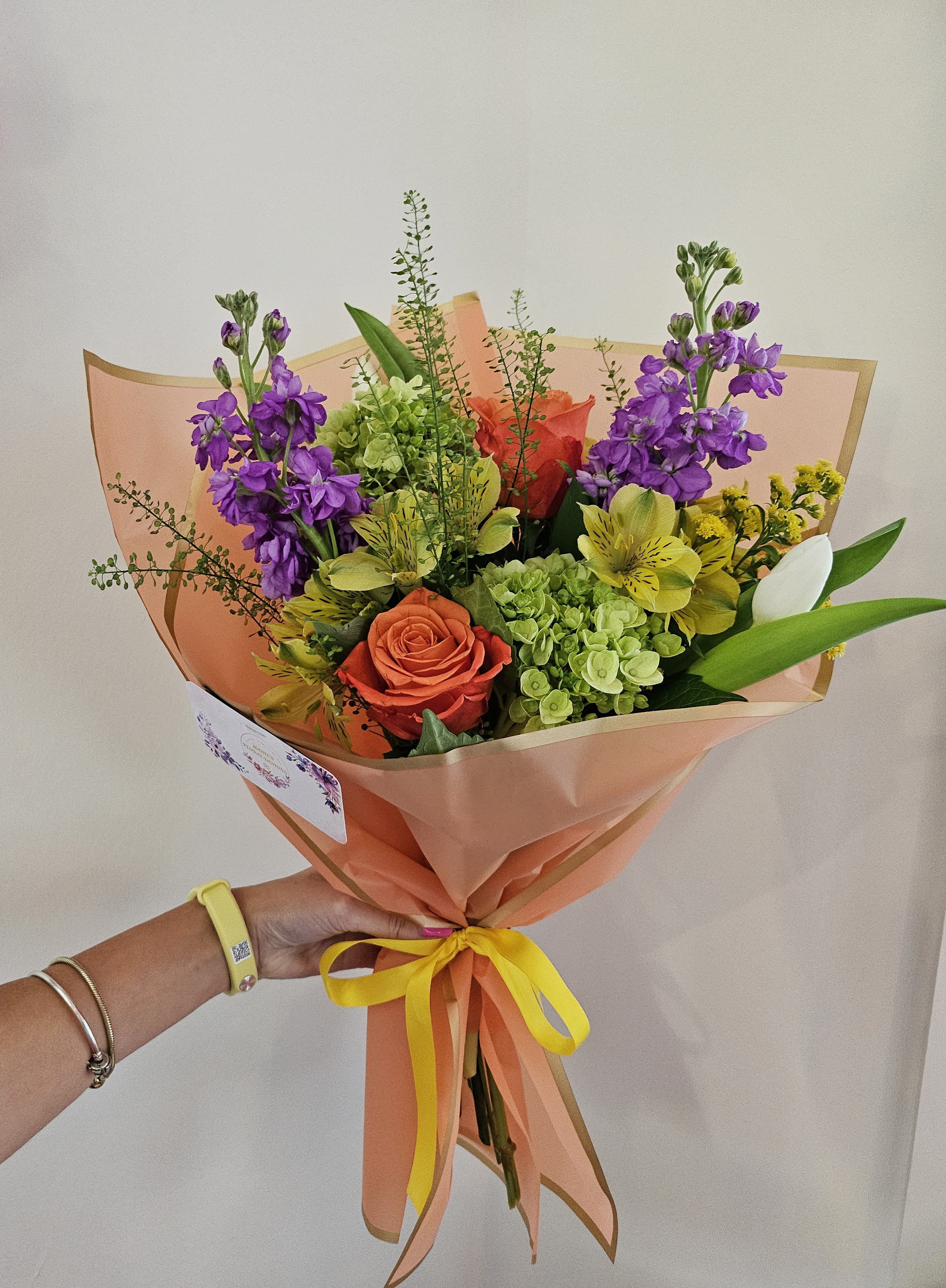 Handheld bouquet of purple, orange, yellow, and green flowers wrapped in peach paper