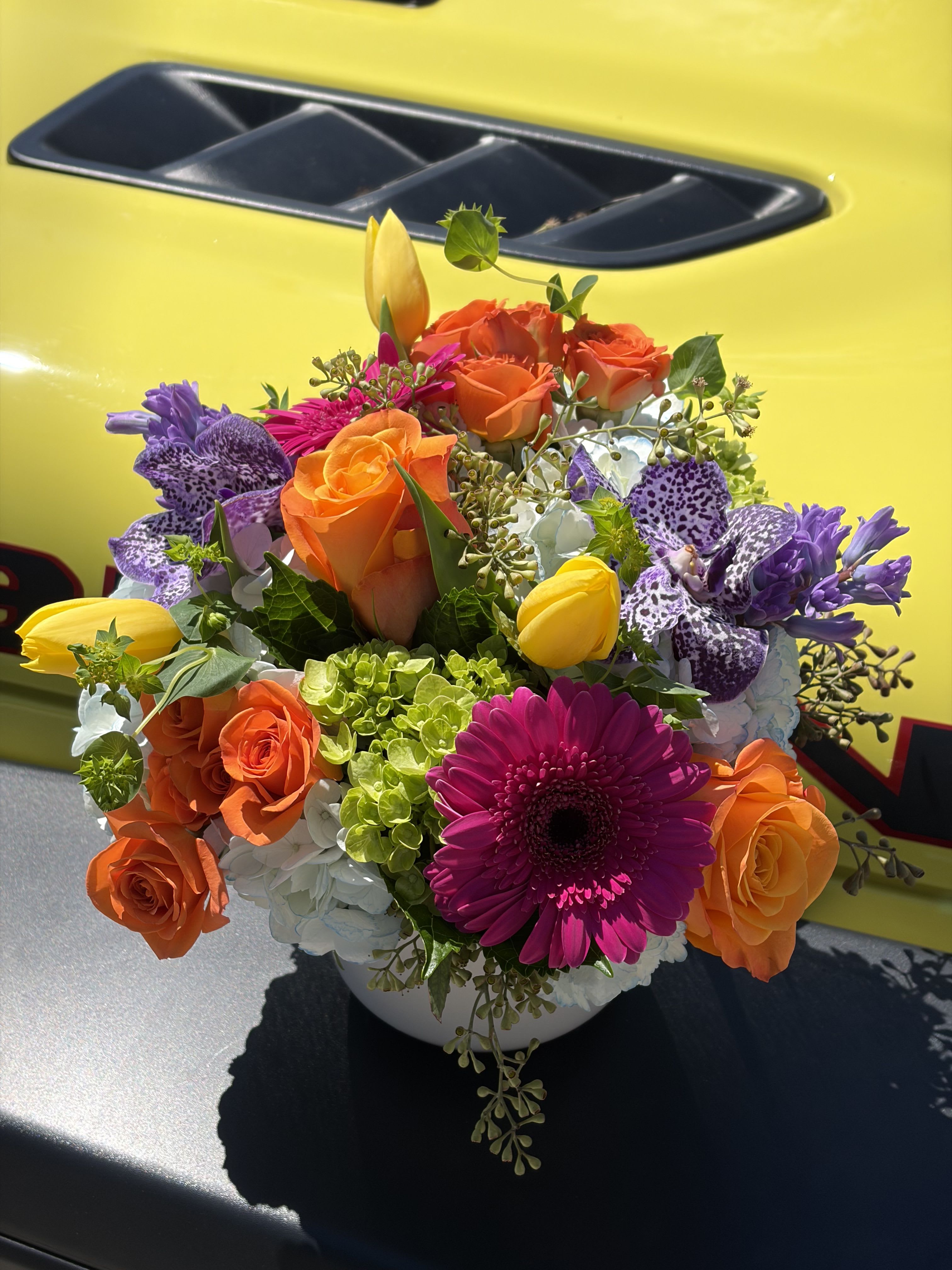 Day Dreaming NY - A vivid celebration in bloom, Day Dreaming is a mixed-color bouquet built to make someone feel truly seen - perfect as a birthday bouquet, congratulations arrangement, or a bright just‑because gift. The arrangement's compact, rounded silhouette lets vivid orange roses, a magenta gerbera daisy, and sunny yellow tulips sit front and center, while purple spotted blooms and purple orchids add unexpected depth for an exuberant, modern look.  Clusters of pale blue and white hydrangea fill the design for a lush, full texture that contrasts beautifully with the sleek petals of tulips and roses. All stems arrive arranged tightly in a simple white ceramic vase that keeps the composition polished and gift-ready; the vase's clean line makes the saturated warm and cool tones pop and suits both casual and more formal settings.  Give this to the person you want to brighten today - it reads as joyful and energetic, the kind of bouquet that elevates a celebration or turns an ordinary afternoon into a moment. Size and stem count are arranged to create a dense, abundant presentation; no special care is needed beyond fresh water and a cool spot away from direct sun to keep blooms vibrant.